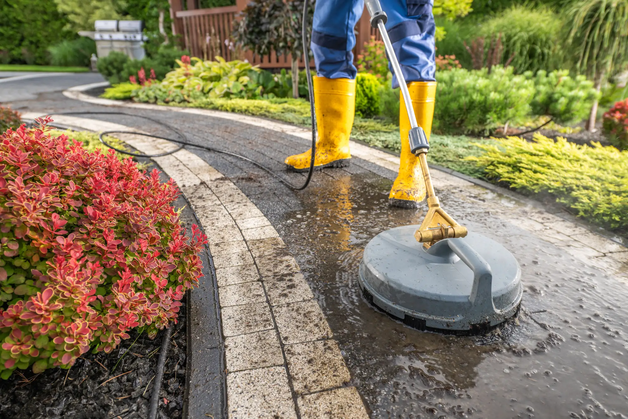 Yellow-booted pro running a surface cleaner over paver bricks — washed pavers bright, unwashed still dark